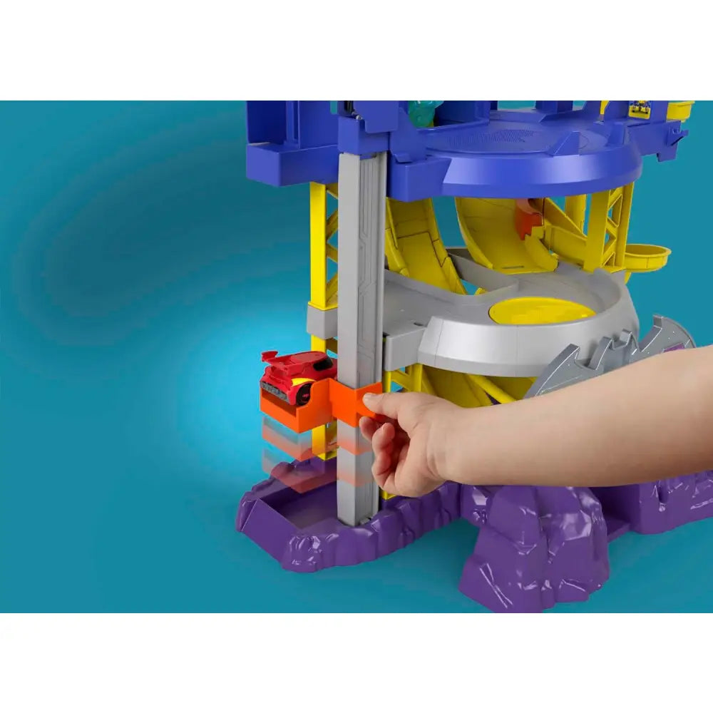 Una mano tiene un’auto hot wheels rosso e arancione vicino all’entrata del playset fisher-price dc batwheels batcaverna lancia e sfreccia